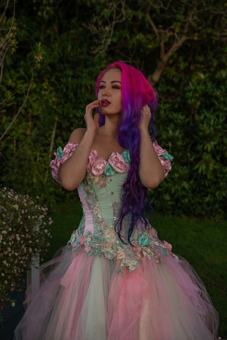 Puede incluir: Una mujer con cabello rosa y morado lleva un cors&eacute; verde claro y rosa con adornos florales y una falda de tul a juego.