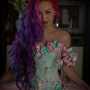 Puede incluir: Una mujer con el pelo largo, morado y rosa lleva un cors&eacute; azul claro con bordados florales rosas y verdes. El cors&eacute; tiene un escote de coraz&oacute;n y se lleva sobre una falda de tul rosa y blanca.
