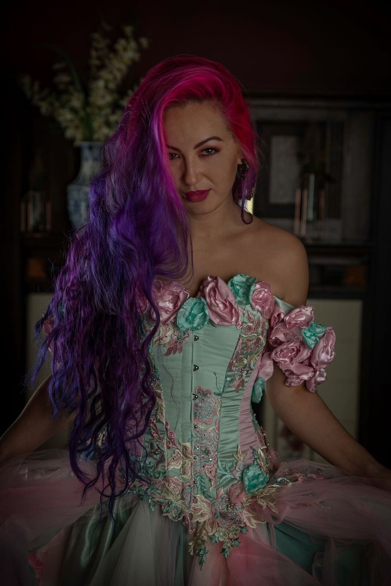 Puede incluir: Una mujer con el pelo largo, morado y rosa lleva un cors&eacute; azul claro con bordados florales rosas y verdes. El cors&eacute; tiene un escote de coraz&oacute;n y se lleva sobre una falda de tul rosa y blanca.