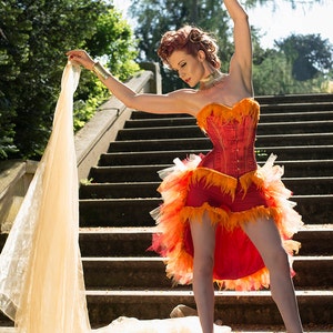 May include: A woman in a red and orange corset with feather trim and a tulle skirt poses on a set of stone stairs. She is holding a sheer fabric in her hands and is wearing a gold necklace and bracelet.