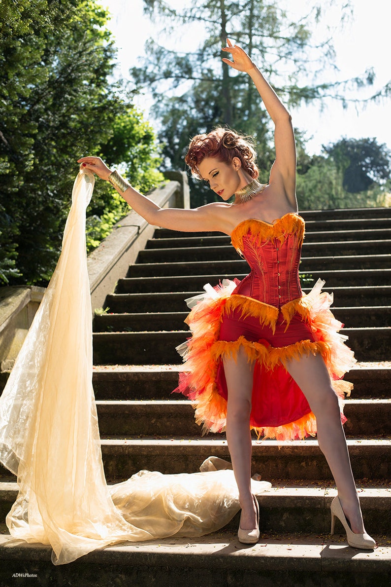 May include: A woman in a red and orange corset with feather trim and a tulle skirt poses on a set of stone stairs. She is holding a sheer fabric in her hands and is wearing a gold necklace and bracelet.