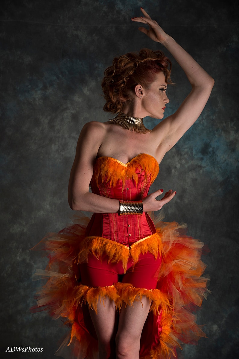 May include: A woman wearing a red corset with orange feathers and a matching skirt. She is wearing a gold choker necklace and a gold bracelet. The background is a dark blue.