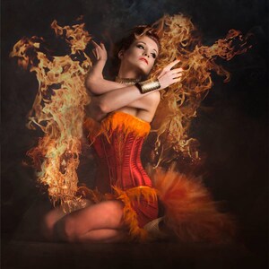 May include: A woman wearing a red and orange corset with feathers, sitting in a pose with flames behind her.