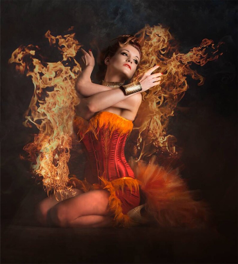 May include: A woman wearing a red and orange corset with feathers, sitting in a pose with flames behind her.