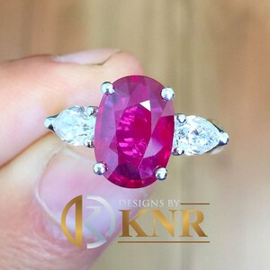 Può includere: Un anello con un grande rubino ovale circondato da due diamanti a forma di pera. L'anello è in oro bianco.