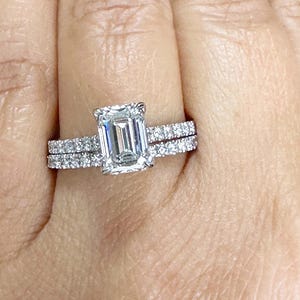 May include: A close-up of a woman's hand wearing a diamond engagement ring with a square-shaped center stone and a matching wedding band with smaller diamonds.