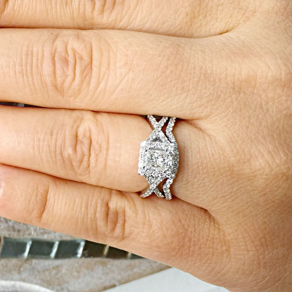 Könnte beinhalten: Nahaufnahme eines Diamantrings mit einem quadratischen Mittelstein und einem Halo aus kleineren Diamanten. Der Ring hat ein einzigartiges Kreuz-Design am Band. Die Hand hat roten Nagellack.