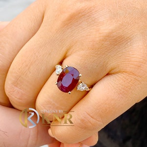 May include: A gold ring with a large oval ruby gemstone and two smaller diamond accents. The ring is on a person's finger.
