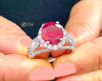 May include: A white gold ring with a large oval ruby surrounded by a halo of smaller diamonds. The ring has a split shank with additional diamonds on each side.