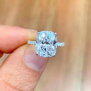 May include: A silver ring with a large, clear, cushion-cut diamond. The diamond is set in a pave setting with smaller diamonds around the center stone.