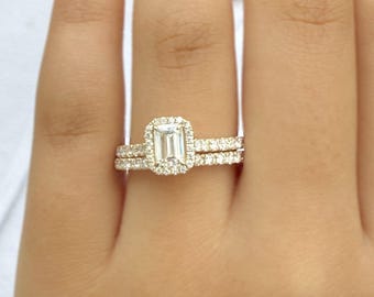 Op de afbeelding: Een diamanten verlovingsringset met een smaragdgeslepen middensteen, omgeven door een halo van kleinere diamanten. De band is ook versierd met diamanten. De ring zit aan een vinger.