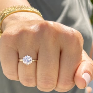 Puede incluir: Primer plano de una mano con un anillo de oro con un gran solitario redondo de diamante transparente.