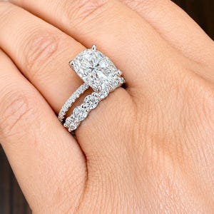 May include: A silver-toned ring set featuring a large cushion-cut center stone held by four prongs on a pavé band, paired with a matching eternity band consisting of multiple round-cut stones set in a continuous row along the metal band.