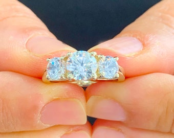 Può includere: Un anello con fascia in oro con tre pietre preziose rotonde e trasparenti. La pietra centrale è più grande delle due pietre su entrambi i lati.