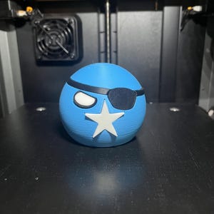 May include: A 3D-printed blue sphere with a white star and eye patch. The sphere has a white eye and a black eye patch. The object is sitting on a black surface.