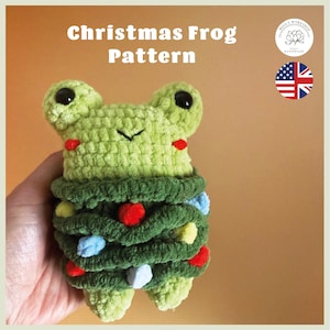 May include: A light green crocheted frog plushie wrapped in a dark green yarn garland decorated with small red, yellow, and light blue pom-poms. The text Christmas Frog Pattern is visible at the top, along with a logo and a US flag icon.