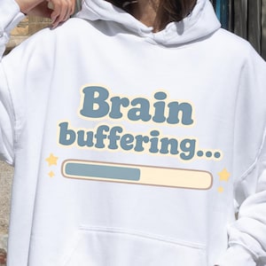 Pode incluir: Moletom branco com a frase "Brain buffering..." em azul e amarelo. O design inclui um gráfico de barra de carregamento. O moletom tem um bolso frontal e um capuz com cordão.