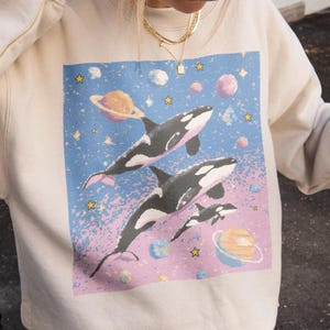 May include: A beige sweatshirt with a graphic of orcas in space, a black purse, and a brown baseball cap with "La Petite Paris" text. The orca graphic has a blue, pink, and purple background with planets. The cap is brown.