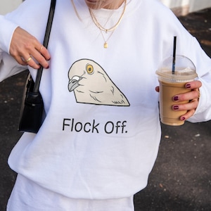 May include: A person wearing a brown baseball cap with "Le Pedre Paris" text, a white sweatshirt with a pigeon graphic and the text "Flock Off.", and a white skirt. The person is holding a black purse and a coffee drink.