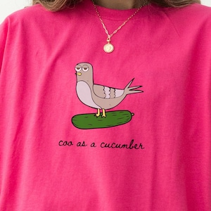 May include: A bright pink t-shirt featuring a cartoon pigeon wearing glasses, perched on a cucumber. The text "coo as a cucumber" is printed below the image. The shirt has short sleeves and a relaxed fit.