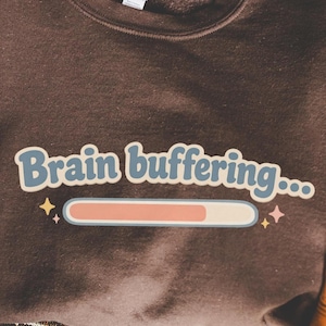 Pode incluir: Um moletom marrom com a frase "Brain buffering..." e uma barra de carregamento. A peça de roupa está dobrada e é segurada por mãos, sobre um fundo estampado.