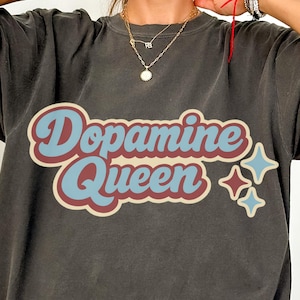 Pode incluir: T-shirt cinza escuro com o texto "Dopamine Queen" em uma fonte retrô. O texto é azul e vermelho, com dois gráficos de estrelas azuis. A camisa tem mangas curtas e um corte folgado.