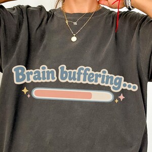 Pode incluir: T-shirt cinzenta escura oversized com o texto "Brain buffering..." em azul claro e um gráfico de barra de carregamento rosa. A t-shirt de manga curta tem um corte folgado, ideal para uso diário.