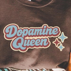 Pode incluir: Um moletom marrom com o texto "Dopamine Queen" em uma fonte retrô, com detalhes em azul e vermelho. Dois gráficos de estrela azul estão no lado direito do texto. O moletom é segurado por duas mãos.