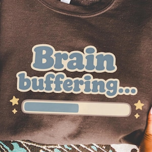 Pode incluir: Um moletom marrom com o texto "Brain buffering..." em azul e branco sujo. O design inclui uma barra de carregamento e estrelas. O moletom é segurado por uma pessoa.