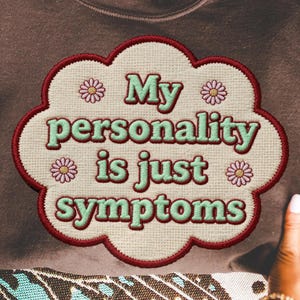 May include: A brown sweatshirt with the embroidered text "My personality is just symptoms" in a cloud-shaped design. The design features pink flowers and a red border. The sweatshirt is held by two hands.