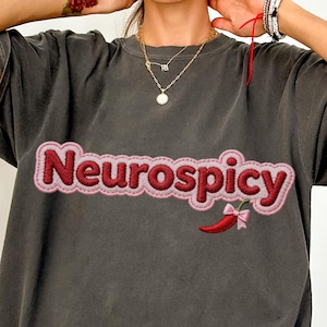 Puede incluir: Camiseta gris oscuro con la palabra "Neurospicy" bordada en rosa y rojo. Un chile rojo con un lazo rosa está bordado debajo del texto. La camiseta tiene mangas cortas y un corte holgado.
