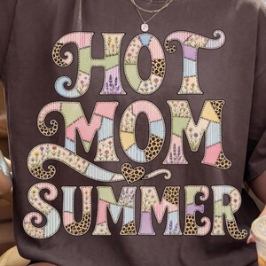 May include: A dark brown short-sleeve t-shirt featuring the text "HOT MOM SUMMER" in a large, multi-patterned patchwork font. The letters incorporate floral, leopard print, and striped designs in pastel colors, with a small heart icon below the word MOM.