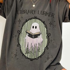 May include: Dark gray t-shirt with the text "LIBRARY LURKER" above an oval frame. Inside the frame is a ghost reading a book. The ghost is white with a pinkish hue, and the book is black.