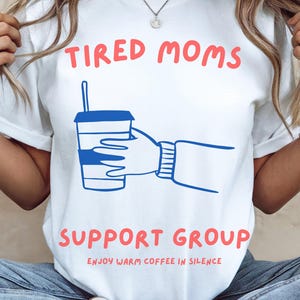 Puede incluir: Una camiseta blanca con el texto en rojo TIRED MOMS SUPPORT GROUP y el texto más pequeño ENJOY WARM COFFEE IN SILENCE. El gráfico central muestra un dibujo lineal azul de una mano sosteniendo un vaso de café con pajita, en estilo simple.