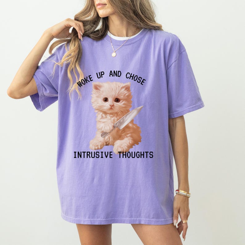 May include: Purple oversized t-shirt featuring a graphic of a cat holding a knife. The text on the shirt reads "Woke up and chose intrusive thoughts".