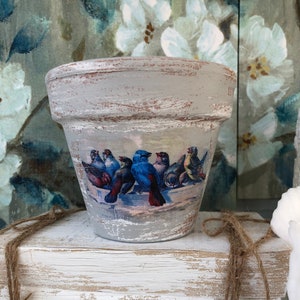 May include: A white ceramic flower pot with a distressed finish and a vintage design of blue birds. The pot is sitting on a white wooden block with twine wrapped around it.