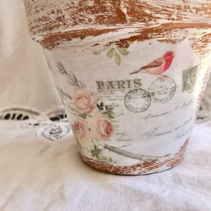 May include: A white terracotta flower pot with a distressed finish. The pot is decorated with a vintage-style design featuring roses, a bird, and the word "PARIS".