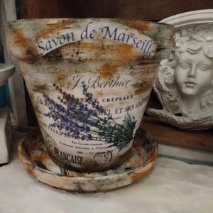 May include: A tapered ceramic flower pot with a matching saucer, featuring a distressed finish in shades of cream, brown, and orange. The pot displays a lavender graphic and black text reading Savon de Marseille and J. Berthier.
