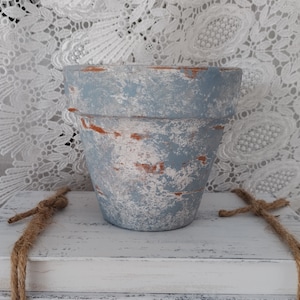 May include: A distressed, light blue and white painted terracotta flower pot. The pot has a weathered appearance with exposed orange clay peeking through the paint. The pot sits on a white surface with a decorative rope.