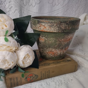 May include: A weathered terracotta flower pot with green and orange paint sits atop a vintage book. Cream-colored faux peonies and green leaves are arranged to the left. The pot has a rustic, aged appearance, perfect for a farmhouse aesthetic.