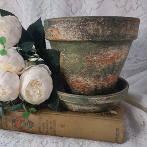May include: A distressed green and orange terracotta flower pot with a matching saucer. The pot sits atop a vintage book and is next to a bouquet of white peonies. The pot has a weathered, aged appearance.