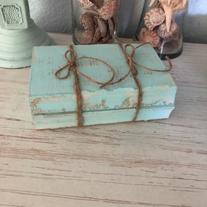 May include: Two stacked books with a light blue painted finish and a rustic, distressed look. The books are tied together with twine and have a vintage feel.