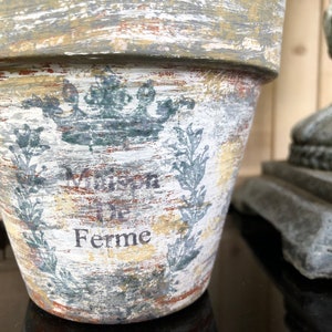 May include: A white ceramic flower pot with a distressed finish. The pot has a crown design and the words "Maison de Ferme" written on it.