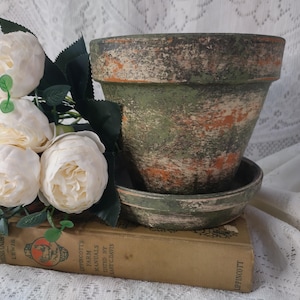 May include: A distressed green and orange ceramic flower pot with a matching saucer. The pot is sitting on top of a vintage book and next to a bouquet of white flowers.