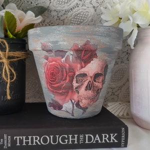 May include: A gray terracotta planter with a distressed finish features a skull and red roses design. The planter is sitting on a black book with the title "Through the Dark" printed on the spine.