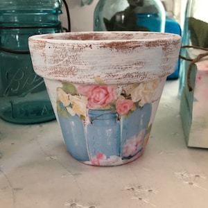 May include: A white terracotta flower pot with a distressed finish. The pot is decorated with a decoupage design of blue mason jars with pink and white flowers.