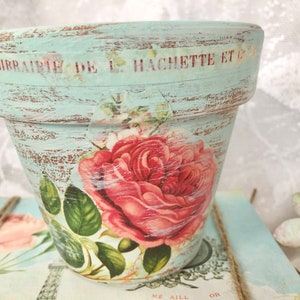 Puede incluir: Una maceta de terracota azul claro con un acabado desgastado. La maceta está decorada con un diseño floral vintage con una rosa rosa y hojas verdes. El texto "Librairie de L. Hachette et C" es visible en el borde superior de la maceta.
