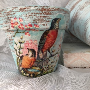 May include: A blue terracotta flower pot with a distressed finish. The pot is decorated with a floral design and two robins.
