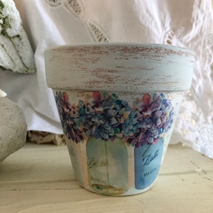 May include: A light blue terracotta flower pot with a distressed finish. The pot is decorated with a floral design and the words "Ball Mason" on the front.