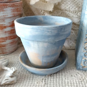 May include: A terracotta plant pot with a matching saucer, featuring a hand-painted finish with blended light blue and white brushstrokes over a natural tan base, showcasing a wide rim and a tapered body design for indoor plant display.